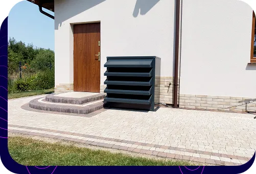 Nowoczesna, grafitowa obudowa akustyczna pompy ciepła, estetycznie wkomponowana w otoczenie domu jednorodzinnego przy wejściu.