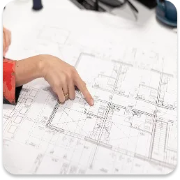 Dłoń wskazująca na fragment dokumentacji projektowej lub planów architektonicznych, co symbolizuje etap analizy i planowania strategii działań przed rozpoczęciem operatu akustycznego