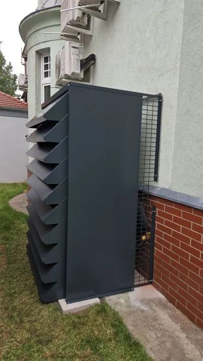 Wysoka, antracytowa obudowa na wymiar, chroniąca dużą pompę ciepła, zamontowana przy ścianie budynku.