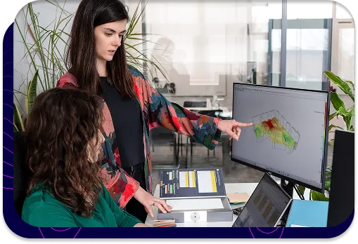 Dwie osoby pracujące w biurze. Jedna z nich wskazuje na monitor komputera, na którym widoczny jest zaawansowany model 3D z analizą akustyczną. To wizualne przedstawienie komputerowego modelowania hałasu, które pomaga w zarządzaniu ryzykiem inwestycyjnym.