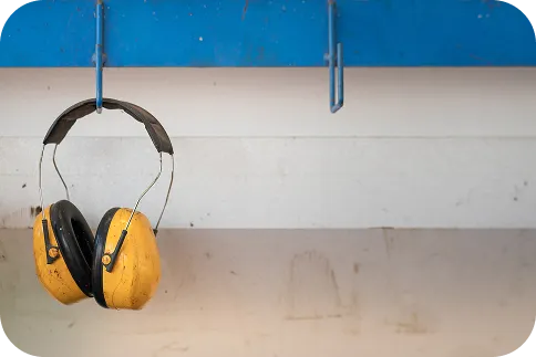 Żółto-czarne słuchawki ochronne, zawieszone na niebieskiej rurze. Obraz symbolizuje ochronę przed hałasem i zarządzanie ryzykiem akustycznym na placu budowy lub rozbiórki, mające na celu minimalizację uciążliwości dla otoczenia.
