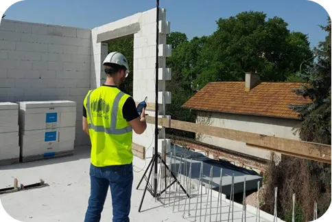 kustyk w kasku i kamizelce ochronnej wykonujący pomiar akustyczny na placu budowy. Obraz ilustruje analizę izolacyjności akustycznej w trakcie realizacji projektu budowlanego,.