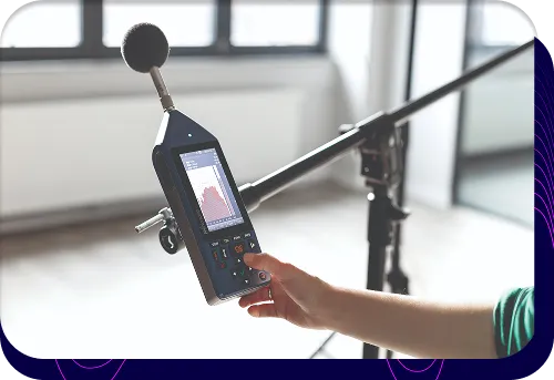 Zbliżenie na profesjonalny miernik poziomu dźwięku, trzymany przez osobę wykonującą pomiar hałasu w pomieszczeniu.