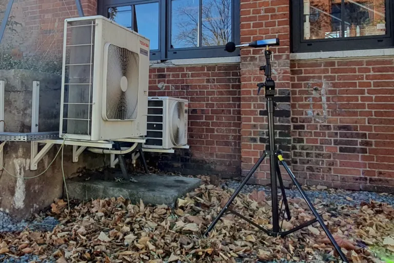 Miernik akustyczny ustawiony na zewnątrz budynku, w pobliżu dwóch jednostek klimatyzacyjnych. Zdjęcie przedstawia proces pomiaru hałasu emitowanego przez urządzenia zewnętrzne.