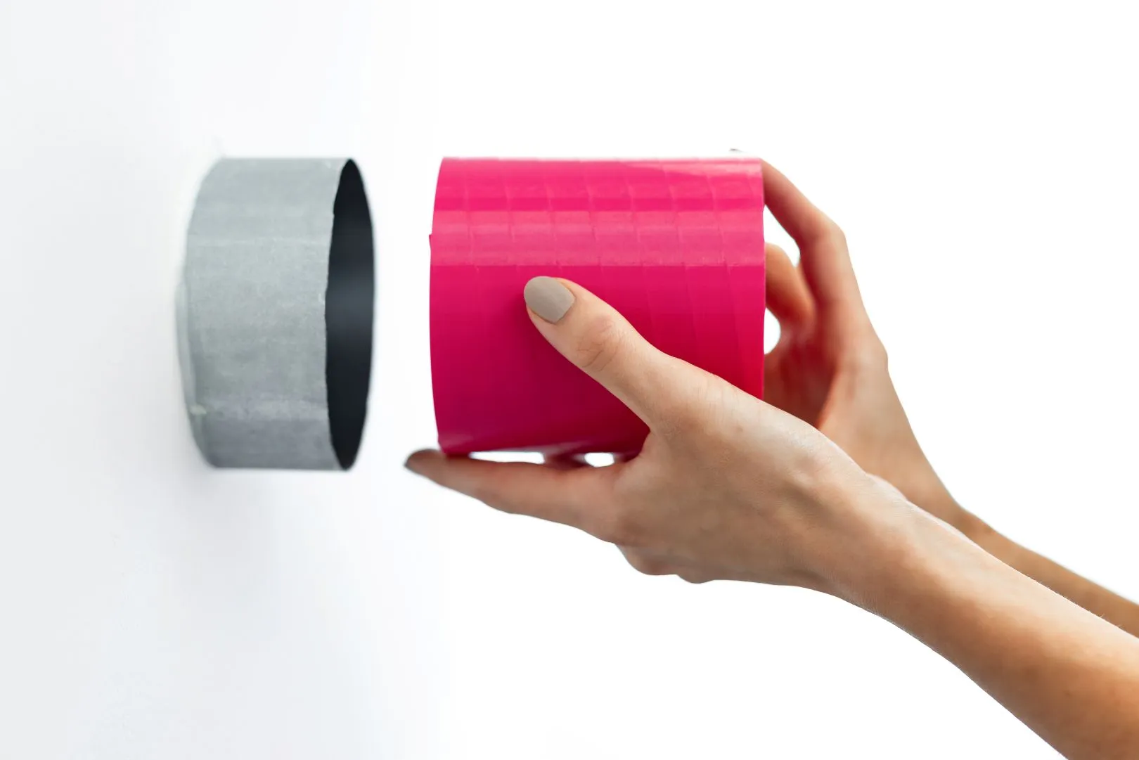 Zdjęcie przedstawia dłonie wsuwające różowy tłumik akustyczny s|TWIST do okrągłego kanału wentylacyjnego w białej ścianie. Scena demonstruje prosty i czysty montaż, idealne dopasowanie produktu oraz łatwość integracji z systemem wentylacji.