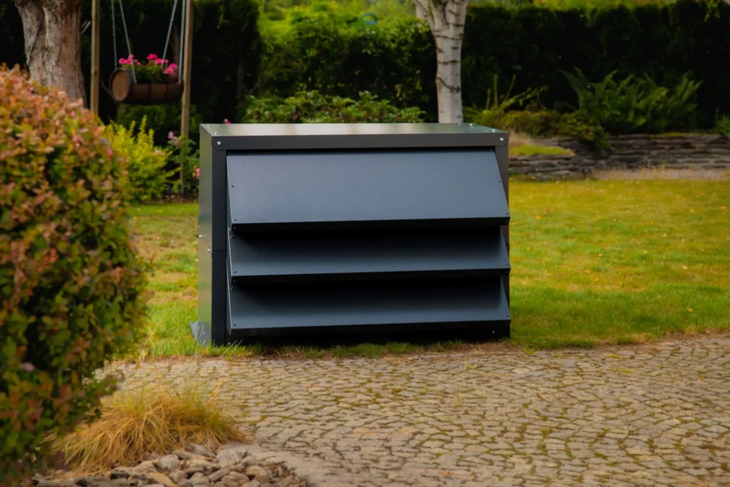 Pompa ciepła w nowoczesnej, grafitowej obudowie akustycznej Silencions, umieszczona na trawniku obok podjazdu z kostki brukowej.