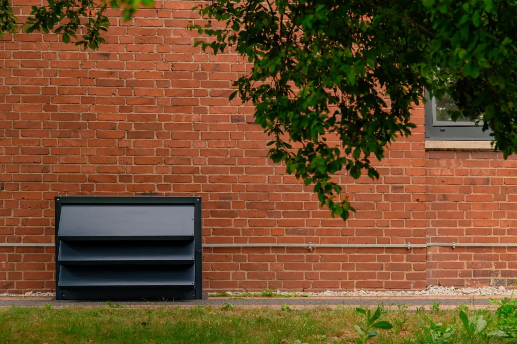 Nowoczesna, grafitowa obudowa akustyczna Silencions do wyciszania pompy ciepła, zainstalowana na zewnątrz przy ścianie domu z cegły.