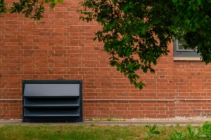 Nowoczesna, grafitowa obudowa akustyczna Silencions do wyciszania pompy ciepła, zainstalowana na zewnątrz przy ścianie domu z cegły.