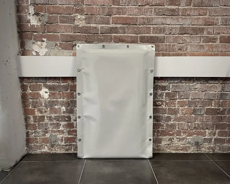 Pionowy, biały panel akustyczny s|FLEX| z widocznymi metalowymi oczkami montażowymi na obwodzie, stojący na tle ceglanej ściany.